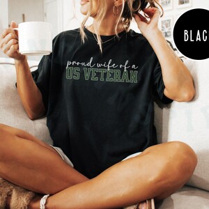 Puede incluir: Camiseta negra con el texto "proud wife of a US VETERAN" en letras blancas y verdes. La camiseta es de corte holgado y la lleva una persona. Una taza blanca se sostiene en la mano de la persona. Un c&iacute;rculo negro con la palabra "BLACK" est&aacute; en la esquina superior derecha.
