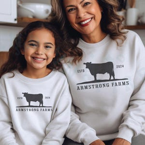 Puede incluir: Dos personas con sudaderas blancas con una silueta de vaca negra y el texto "ESTD 2024 ARMSTRONG FARMS".