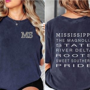 May include: Navy blue t-shirt with the letters "MS" in a cream color on the front. The back of the shirt features the words "Mississippi, The Magnolia State, River Delta, Roots, Sweet Southern Pride" in a vertical layout.