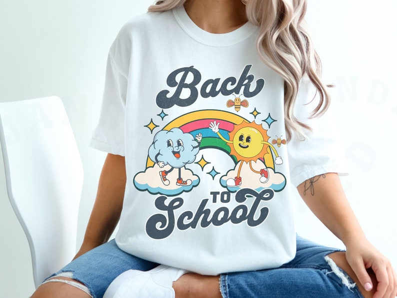 May include: A white t-shirt with a retro-style graphic design featuring a smiling sun and a cloud holding hands over a rainbow. The text 'Back to School' is written in a retro font below the graphic.