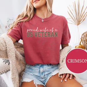 Puede incluir: Camiseta de color carmes&iacute; con el texto "proud wife of a US VETERAN" en fuente de escritura. La camiseta se combina con pantalones cortos de mezclilla. Un c&iacute;rculo redondo de color carmes&iacute; con la palabra "CRIMSON" est&aacute; en la esquina inferior derecha.