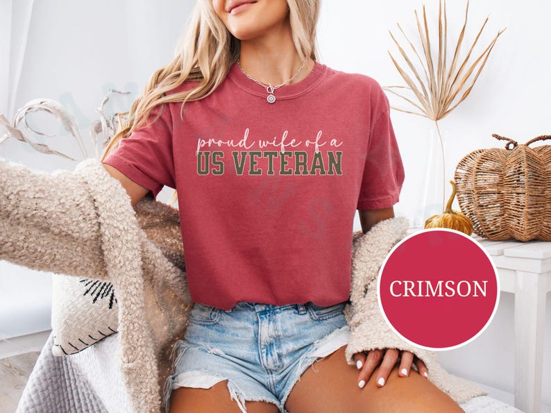 Puede incluir: Camiseta de color carmes&iacute; con el texto "proud wife of a US VETERAN" en fuente de escritura. La camiseta se combina con pantalones cortos de mezclilla. Un c&iacute;rculo redondo de color carmes&iacute; con la palabra "CRIMSON" est&aacute; en la esquina inferior derecha.