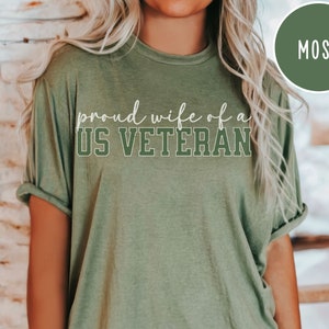 Puede incluir: Camiseta verde musgo con el texto "proud wife of a US VETERAN" en escritura blanca y letras de bloque. La camiseta tiene mangas enrolladas y cuello redondo. Un gr&aacute;fico circular en la esquina superior derecha dice "MOSS".