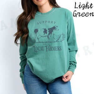 May include: A light green long-sleeved t-shirt with a graphic of a cow and a chicken. The text on the shirt reads "Support Your Local Farmers".