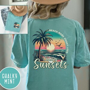 May include: A teal blue t-shirt with a graphic of a palm tree and sunset on the back. The text "Beach More Worry Less" is above the graphic and "Chasing Sunsets" is below the graphic.
