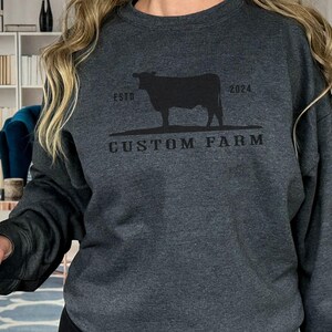 Puede incluir: Una sudadera gris oscuro con una silueta negra de una vaca y el texto "ESTD 2024 CUSTOM FARM".