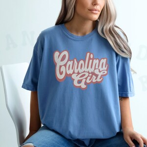 May include: A woman wearing a blue t-shirt with the text "Carolina Girl" in pink and white lettering. The shirt has a vintage style.