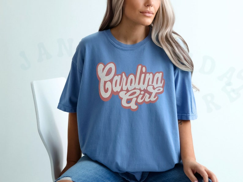 May include: A woman wearing a blue t-shirt with the text "Carolina Girl" in pink and white lettering. The shirt has a vintage style.