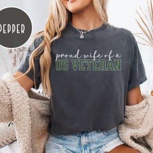 Puede incluir: Camiseta gris oscuro con el texto "proud wife of a US VETERAN" en blanco y verde. La camiseta tiene un aspecto vintage. La modelo lleva una rebeca color crema y unos shorts vaqueros. El color de la camiseta es pimienta.