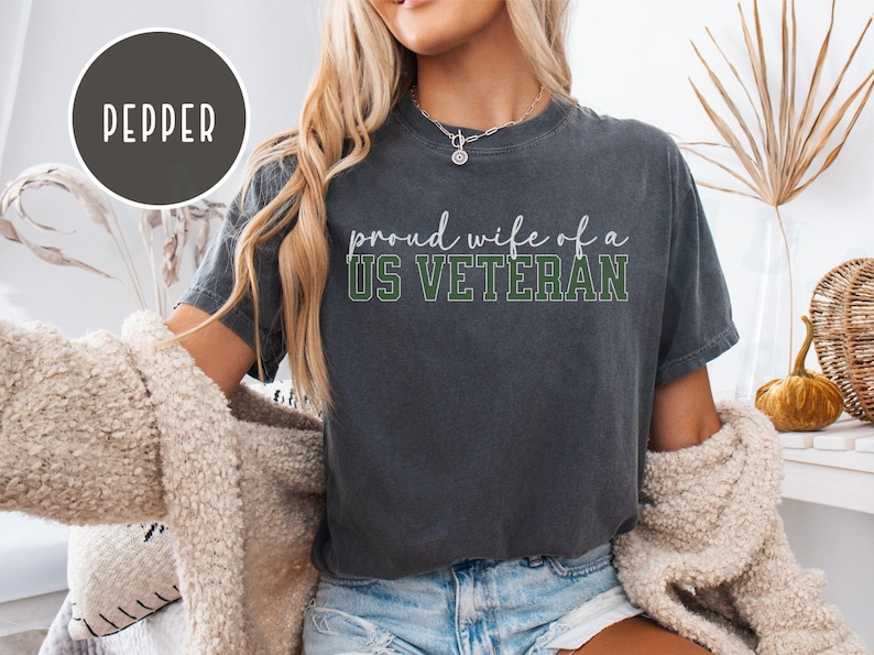 Puede incluir: Camiseta gris oscuro con el texto "proud wife of a US VETERAN" en blanco y verde. La camiseta tiene un aspecto vintage. La modelo lleva una rebeca color crema y unos shorts vaqueros. El color de la camiseta es pimienta.