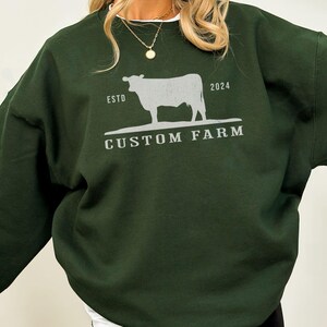 Puede incluir: Una sudadera verde oscuro con una silueta blanca de una vaca y el texto "ESTD 2024 CUSTOM FARM".