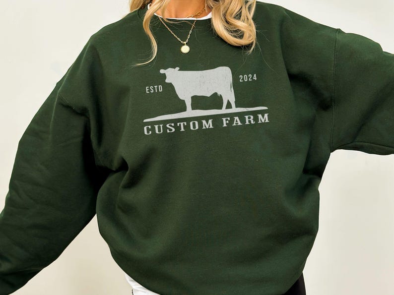 Puede incluir: Una sudadera verde oscuro con una silueta blanca de una vaca y el texto "ESTD 2024 CUSTOM FARM".