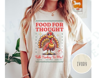 May include: A white t-shirt with a vintage-style graphic of a turkey with the text "Food for Thought" and "Talk Turkey To Me!" in red and green. The shirt is labeled "Ivory".