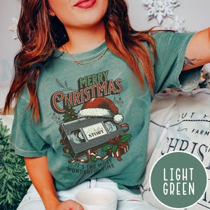 May include: A light green t-shirt with a vintage-style Christmas design featuring a VHS tape with the words "Merry Christmas" and "It's the most wonderful time" printed on it.