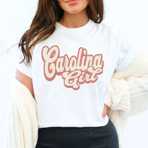 May include: White t-shirt with a pink and cream graphic that says "Carolina Girl" in a retro font.