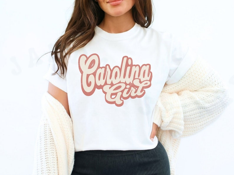 May include: White t-shirt with a pink and cream graphic that says "Carolina Girl" in a retro font.