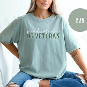 Puede incluir: Camiseta verde claro con el texto "proud wife of a US VETERAN" en letras blancas y verdes. La camiseta tiene un corte holgado y mangas cortas. La modelo lleva vaqueros azules.