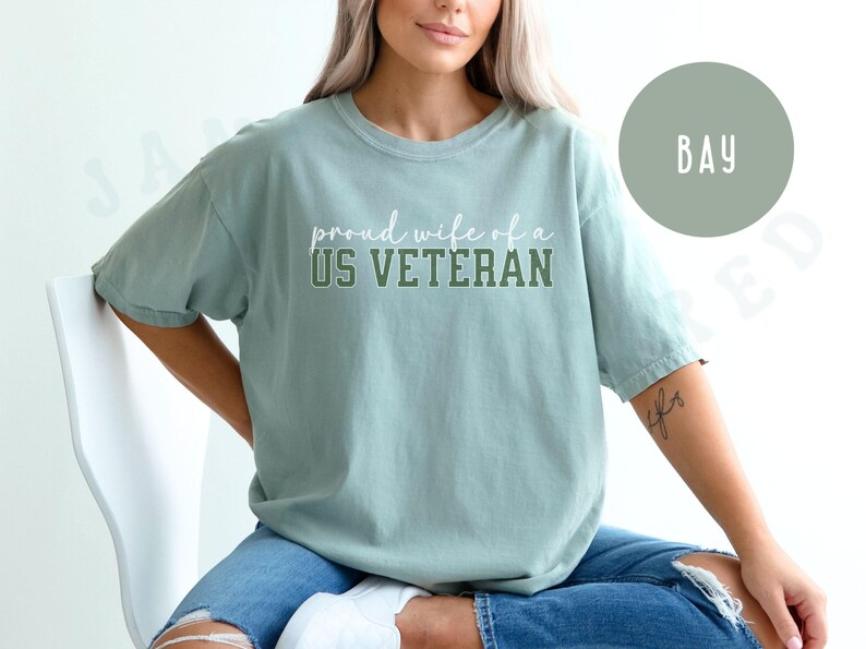 Puede incluir: Camiseta verde claro con el texto "proud wife of a US VETERAN" en letras blancas y verdes. La camiseta tiene un corte holgado y mangas cortas. La modelo lleva vaqueros azules.