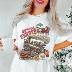 May include: White t-shirt featuring a vintage illustration of a train with the text "North Pole Express." Santa Claus and reindeer are depicted above the train. The design includes the text "Believe in the Magic Powered by Joy Rides."