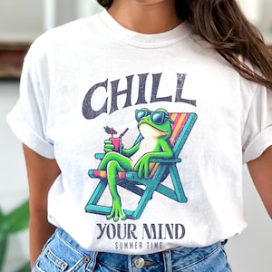 May include: White t-shirt featuring a cartoon frog wearing sunglasses and relaxing in a beach chair, holding a drink. The shirt has the words "CHILL YOUR MIND SUMMER TIME" printed above and below the frog illustration.