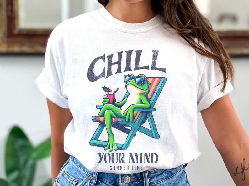 May include: White t-shirt featuring a cartoon frog wearing sunglasses and relaxing in a beach chair, holding a drink. The shirt has the words "CHILL YOUR MIND SUMMER TIME" printed above and below the frog illustration.