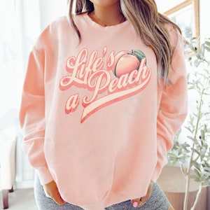 May include: A pink sweatshirt with the text "Life's a Peach" in a retro font. A peach graphic is included in the text.