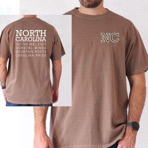 May include: A brown t-shirt with the text "NORTH CAROLINA" on the back, including phrases like "The Tar Heel State" and "Carolina Pride." The front displays the letters "NC" in a green and white outline.