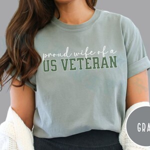 Puede incluir: Camiseta gris con el texto "proud wife of a US VETERAN" en letras blancas y verdes. La camiseta tiene mangas enrolladas y un corte relajado. Un c&iacute;rculo gris con la palabra "GRAY" est&aacute; en la esquina inferior derecha.