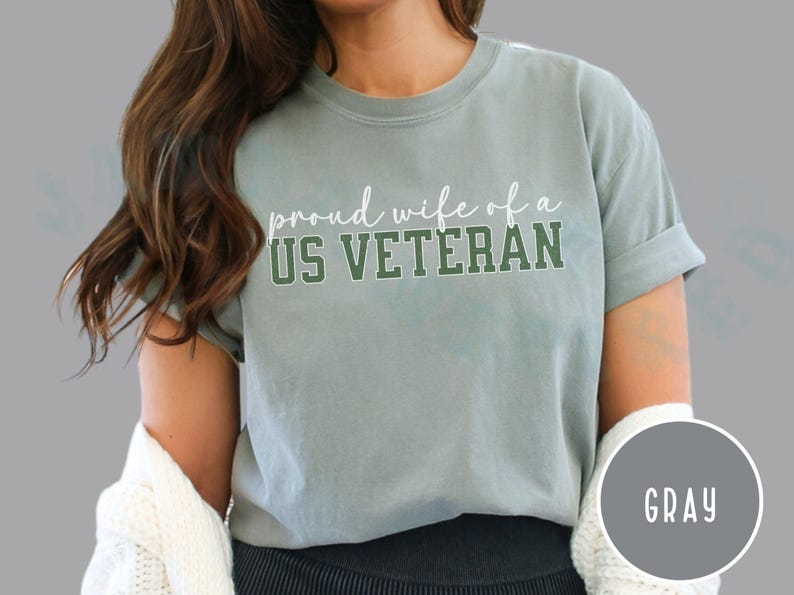 Puede incluir: Camiseta gris con el texto "proud wife of a US VETERAN" en letras blancas y verdes. La camiseta tiene mangas enrolladas y un corte relajado. Un c&iacute;rculo gris con la palabra "GRAY" est&aacute; en la esquina inferior derecha.