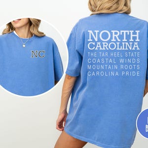 May include: Flo blue t-shirt with "NORTH CAROLINA" printed on the back, along with "THE TAR HEEL STATE", "COASTAL WINDS", "MOUNTAIN ROOTS", and "CAROLINA PRIDE". The front features "NC" in a collegiate style.
