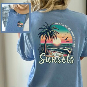 May include: Light blue t-shirt with a graphic of a palm tree, sunset, and ocean waves. The text "Beach More Worry Less" is printed in a circle around the graphic. The text "Chasing Sunsets" is printed below the graphic.