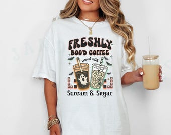 May include: White t-shirt with a Halloween-themed graphic. The design features two coffee cups with spooky illustrations, the text "FRESHLY BOO'D COFFEE" and "Scream & Sugar". A glass of coffee is held in the hand.