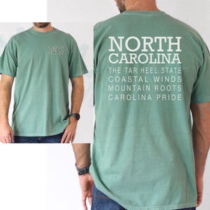 May include: Sage green t-shirt featuring "NC" embroidery on the front and "NORTH CAROLINA" printed on the back, with state-related text. The shirt is short-sleeved and has a crew neck.