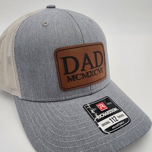 May include: A gray and white trucker hat with a brown leather patch that says "DAD MCMXCVI" on it. The hat is adjustable and has a Richardson logo on the back.