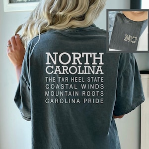 May include: A dark teal t-shirt with the words "NORTH CAROLINA" in large white block letters. Below, smaller text reads "THE TAR HEEL STATE, COASTAL WINDS, MOUNTAIN ROOTS, CAROLINA PRIDE." An inset shows a similar shirt with "NC" on it.