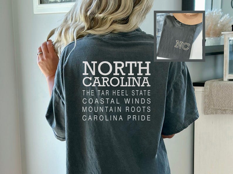 May include: A dark teal t-shirt with the words "NORTH CAROLINA" in large white block letters. Below, smaller text reads "THE TAR HEEL STATE, COASTAL WINDS, MOUNTAIN ROOTS, CAROLINA PRIDE." An inset shows a similar shirt with "NC" on it.
