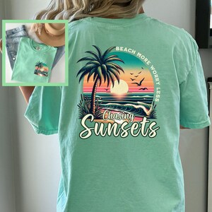 May include: A light green t-shirt with a graphic print of a palm tree, sunset, and ocean waves. The text "Beach More Worry Less" is printed in a circle around the graphic. The text "Chasing Sunsets" is printed below the graphic.