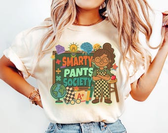 May include: Cream-colored t-shirt featuring a colorful graphic with the text "Smarty Pants Society." The design includes a cartoon illustration of a girl, school supplies, and various educational symbols. The shirt is a casual wear item.