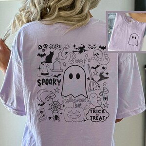 May include: Light purple t-shirt with a black Halloween-themed graphic on the back. The design includes a ghost, bats, a witch's hat, a spiderweb, and the words "Spooky" and "Trick or Treat". A smaller image of the shirt with a ghost is in the corner.