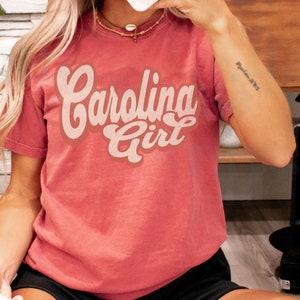 May include: A red t-shirt with the text "Carolina Girl" in a retro font. The text is in a cream colour.