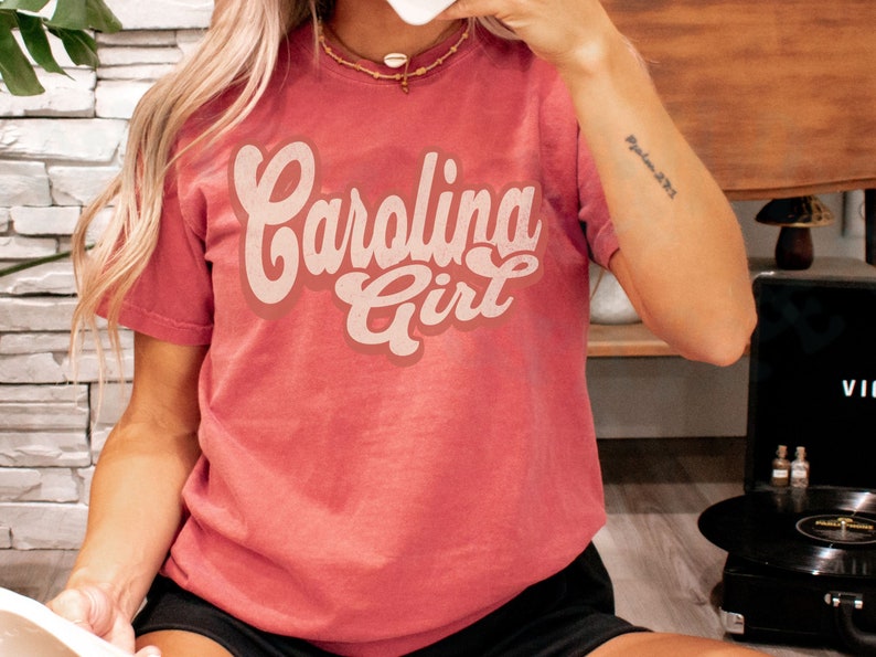 May include: A red t-shirt with the text "Carolina Girl" in a retro font. The text is in a cream colour.