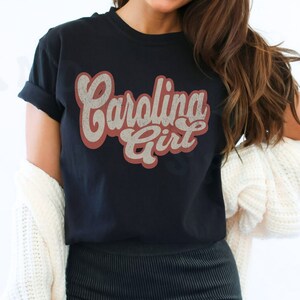 May include: A black t-shirt with a vintage-style graphic that says "Carolina Girl" in red and white lettering.