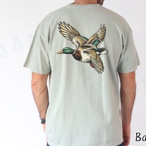 Może przedstawiać: Jasnozielony t-shirt z grafiką lecącego dzikiego kaczora na plecach.