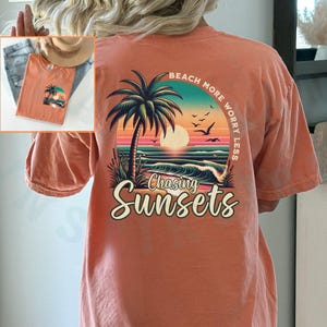 May include: A coral orange t-shirt with a graphic print of a palm tree, sunset, and waves. The text "Beach More Worry Less" is printed in a semi-circle around the graphic. The text "Chasing Sunsets" is printed below the graphic.