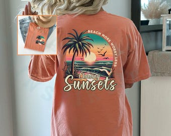 May include: A coral orange t-shirt with a graphic print of a palm tree, sunset, and waves. The text "Beach More Worry Less" is printed in a semi-circle around the graphic. The text "Chasing Sunsets" is printed below the graphic.