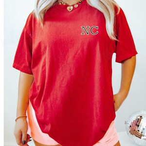 May include: A red t-shirt with the letters "NC" embroidered on the upper left chest. The shirt has short sleeves and a relaxed fit. The person is wearing a gold necklace and bracelet. The background is a light colour.
