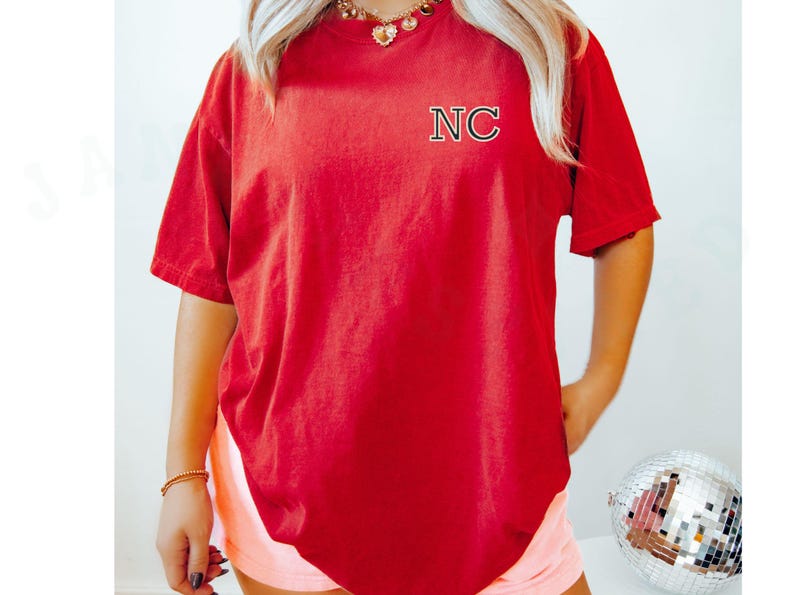 May include: A red t-shirt with the letters "NC" embroidered on the upper left chest. The shirt has short sleeves and a relaxed fit. The person is wearing a gold necklace and bracelet. The background is a light colour.