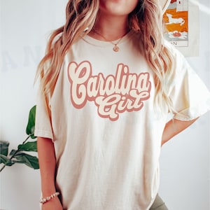 May include: A white t-shirt with a pink and cream graphic that says "Carolina Girl" in a retro font.