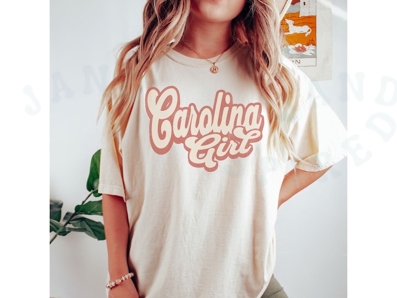 May include: A white t-shirt with a pink and cream graphic that says "Carolina Girl" in a retro font.