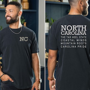 May include: Dark grey t-shirt with "NC" embroidered on the front. The back of the shirt features the words "NORTH CAROLINA" above text that reads "THE TAR HEEL STATE, COASTAL WINDS, MOUNTAIN ROOTS, CAROLINA PRIDE."